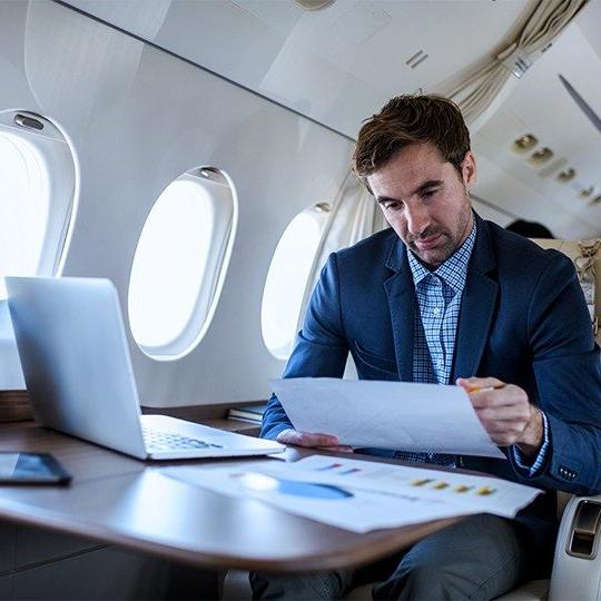 一名男子坐在私人飞机的桌子前，一边用笔记本电脑工作，一边阅读文书