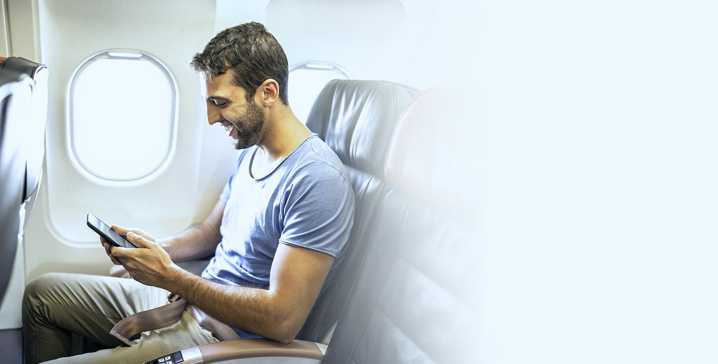 一个留着棕色头发和胡子的男人坐在飞机上靠窗看他的智能手机