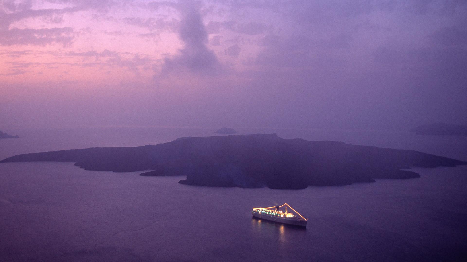 An aerial view of a cruise ship with lights on deck and on the rigging, on the sea in front of a remote island.